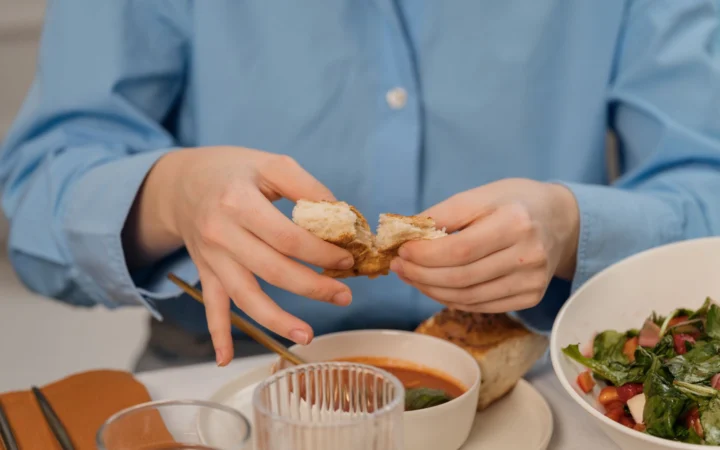Woman tearing bread over a bowl of soup, wearing a blue shirt, indoors.